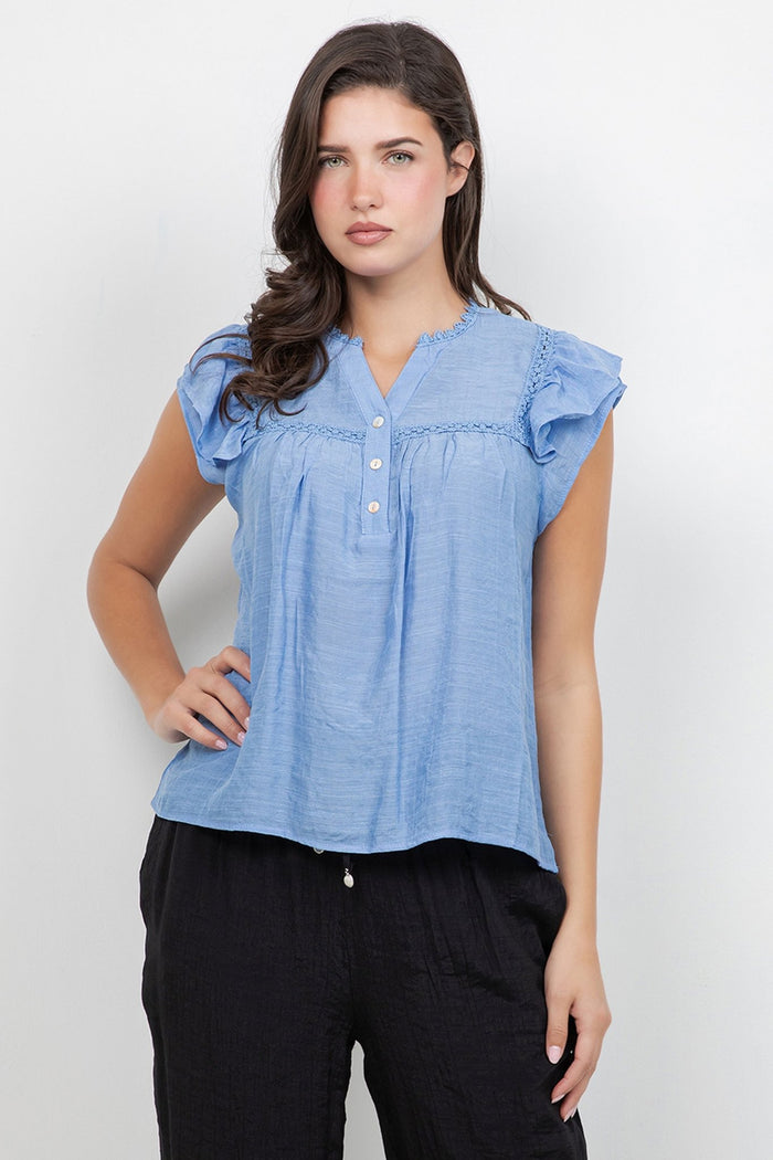 A woman wears a white Ladies Boho Flutter Sleeve Button Blouse and green pants, front view.