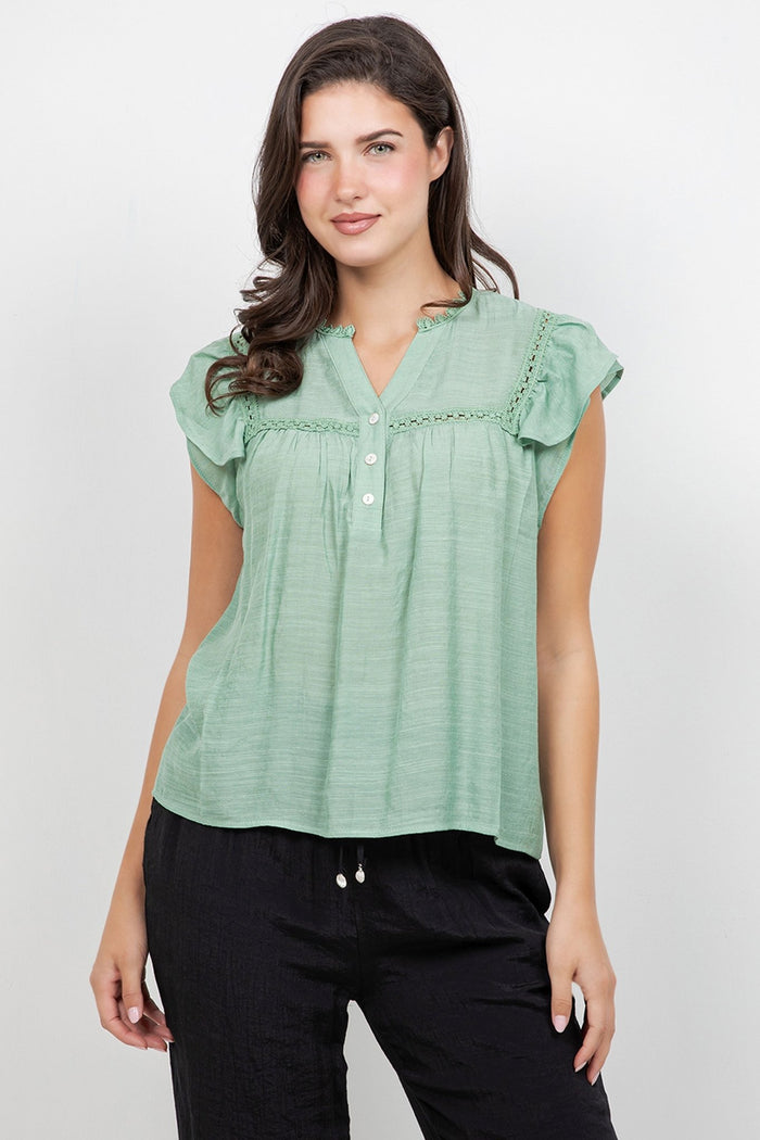 A woman wears a white Ladies Boho Flutter Sleeve Button Blouse and green pants, front view.