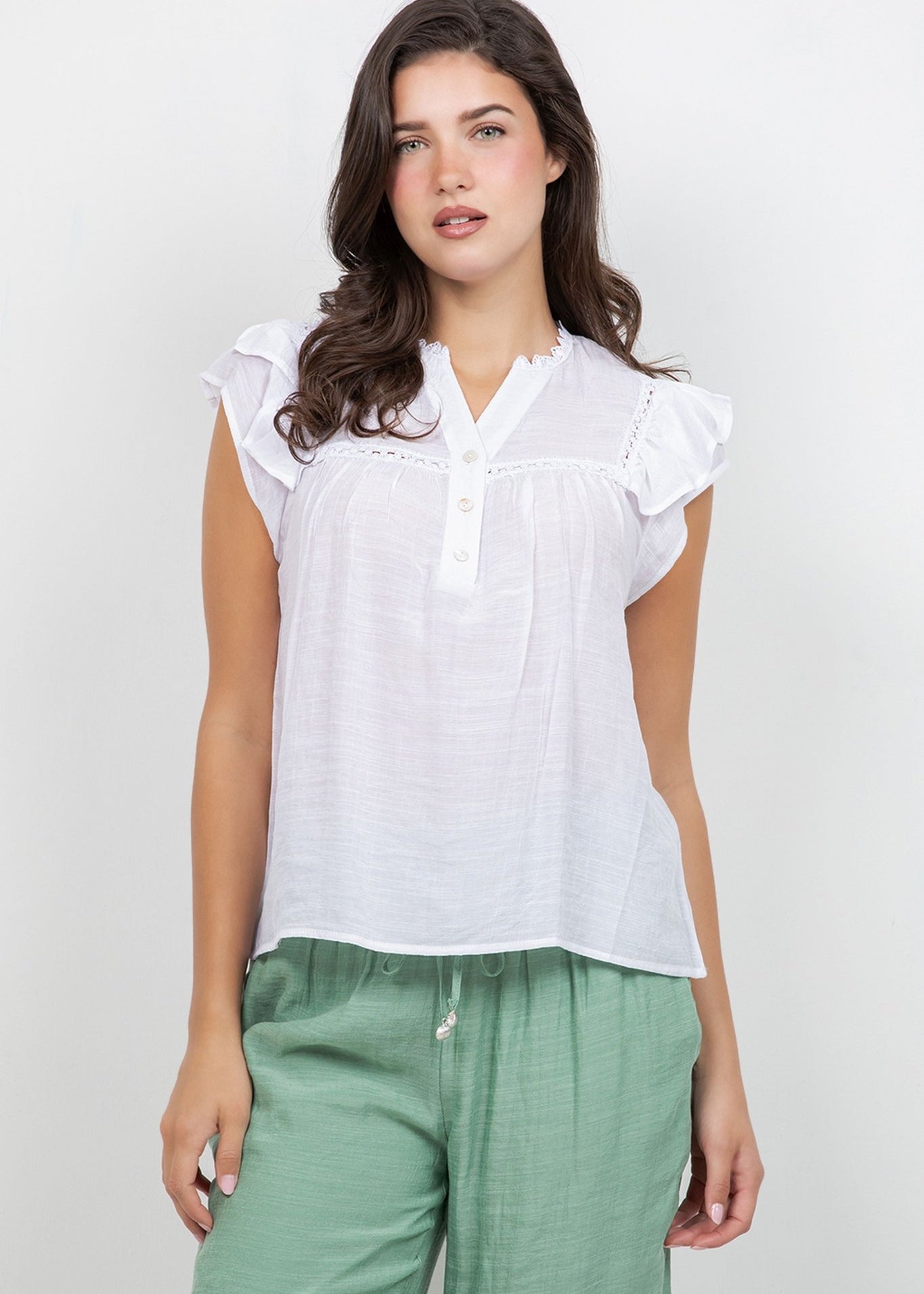 A woman wears a white Ladies Boho Flutter Sleeve Button Blouse and green pants, front view.