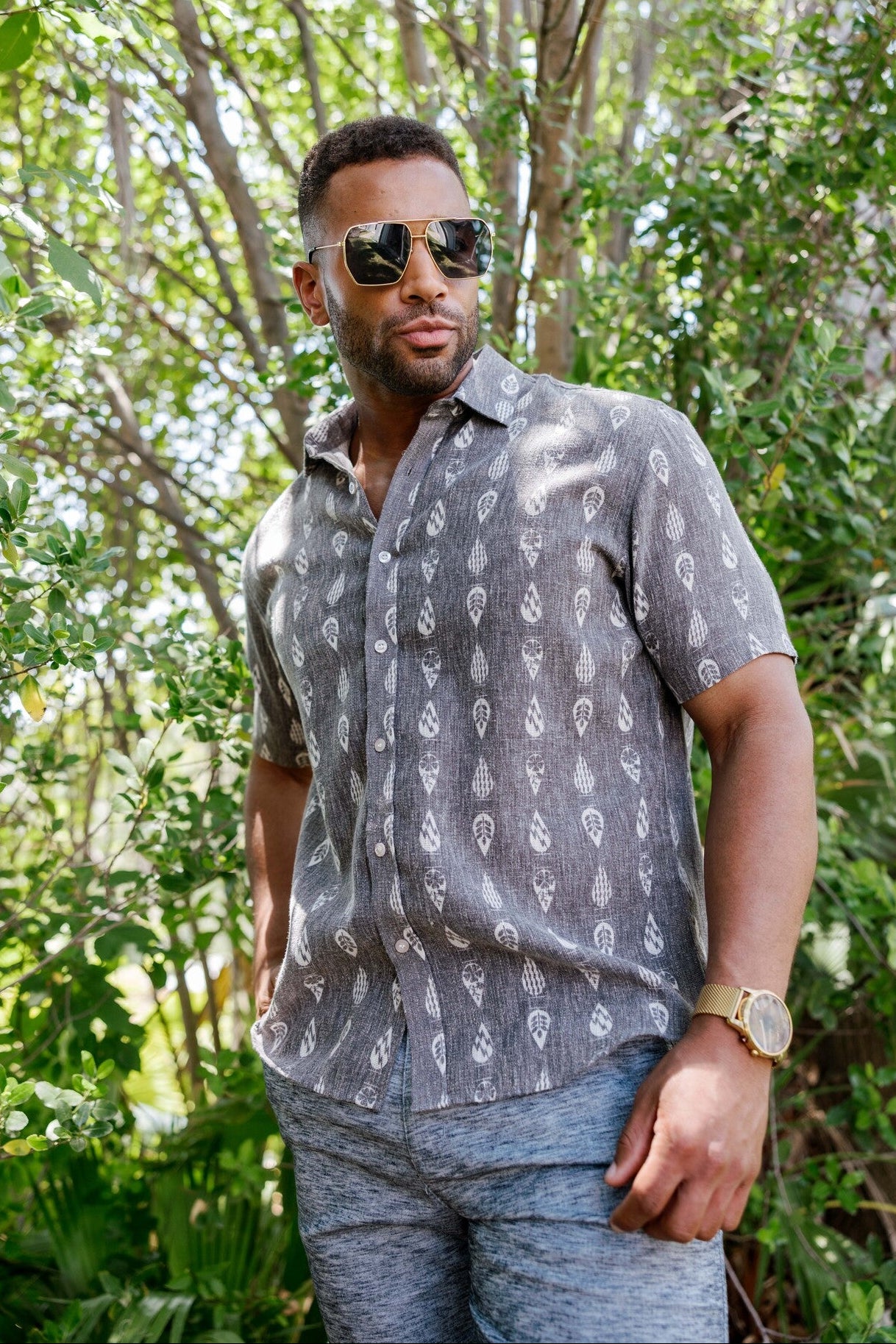 Man wears the gray resort print men's short sleeve button-up shirt outdoors, sunglasses on, looking off to the side.