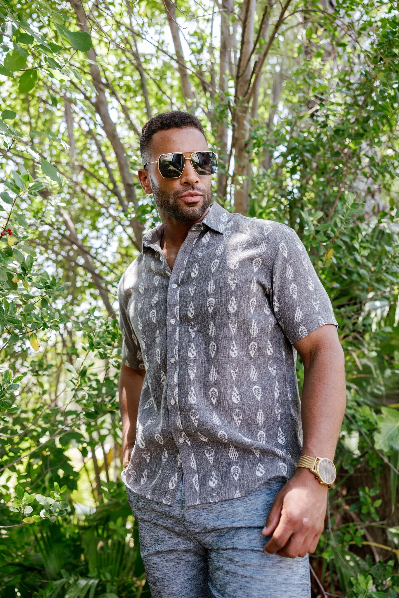 Man wears the gray resort print men's short sleeve button-up shirt outdoors, sunglasses on, looking off to the side.