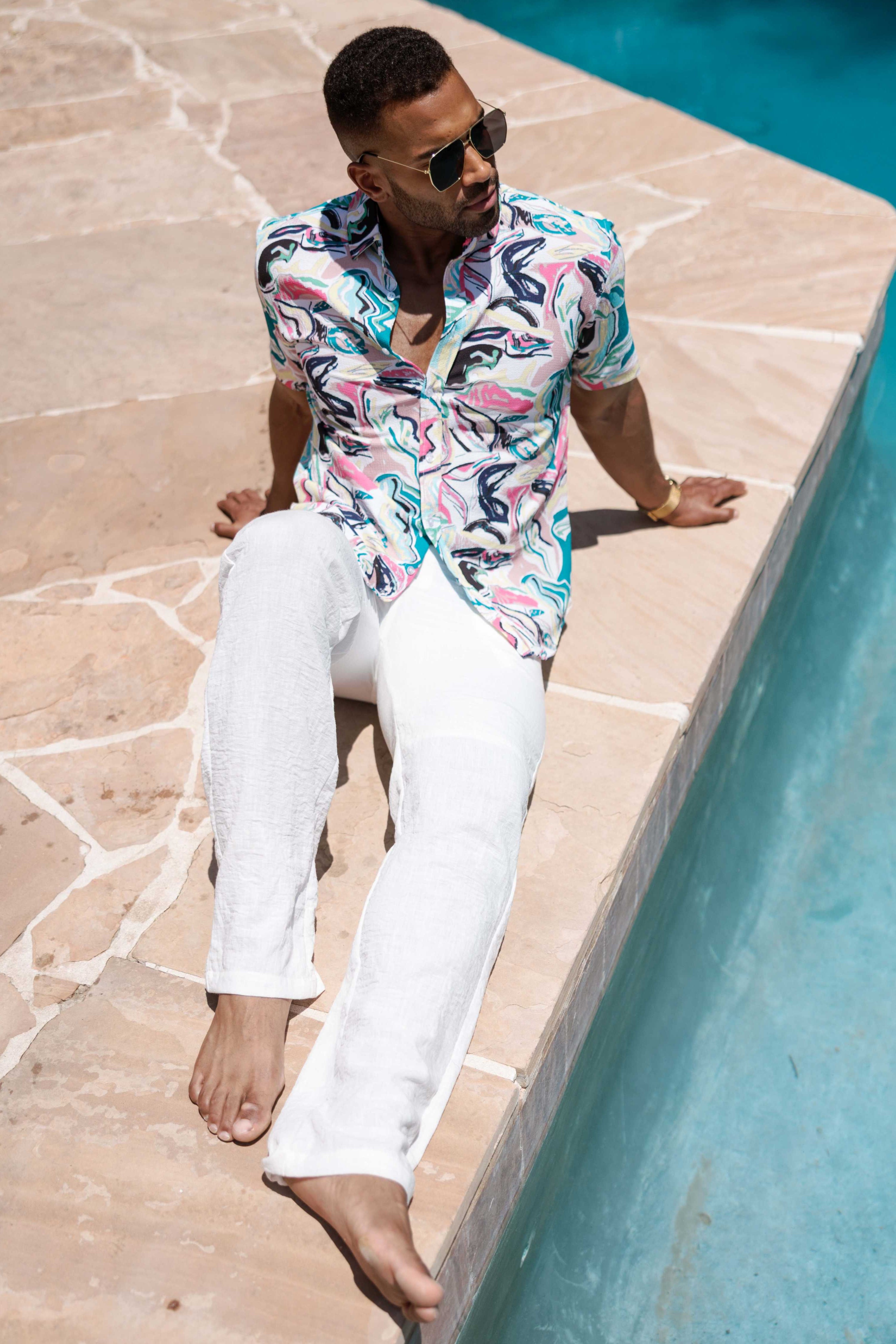 Man sitting by a pool wearing a colorful shirt and white pants.