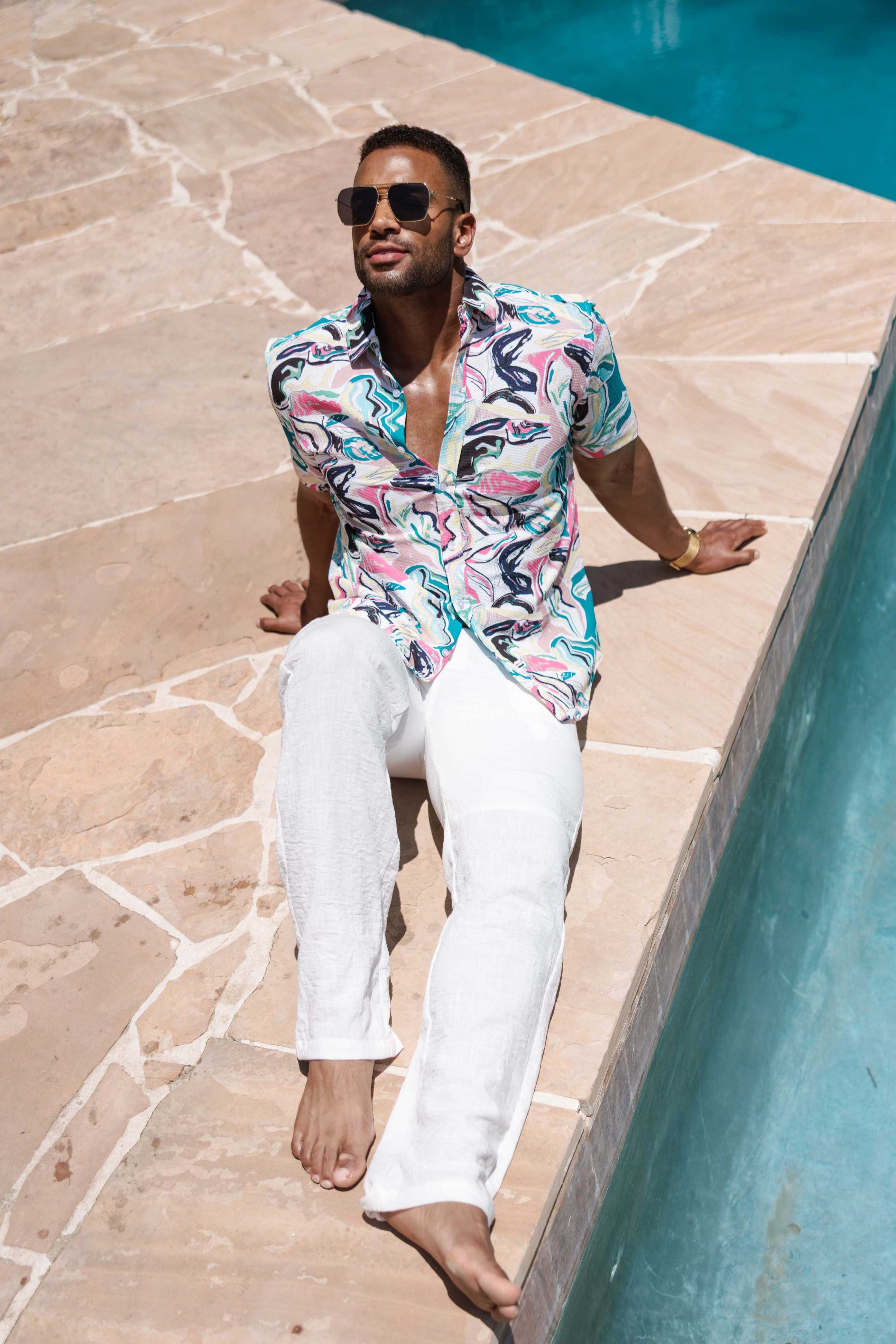 Man in a colorful shirt and white pants sitting by a pool.