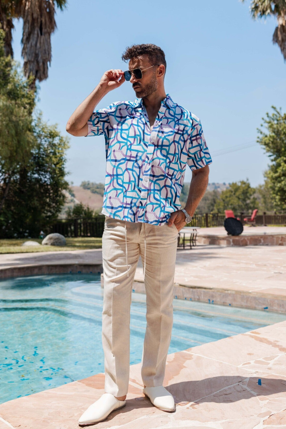 Man in a colorful shirt and beige pants standing by a pool with palm trees in the background