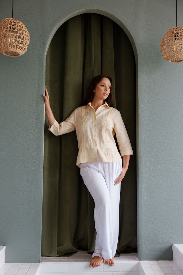 Full body lifestyle shot of a women’s pale yellow linen guayabera paired with white pants for a beach dinner look.