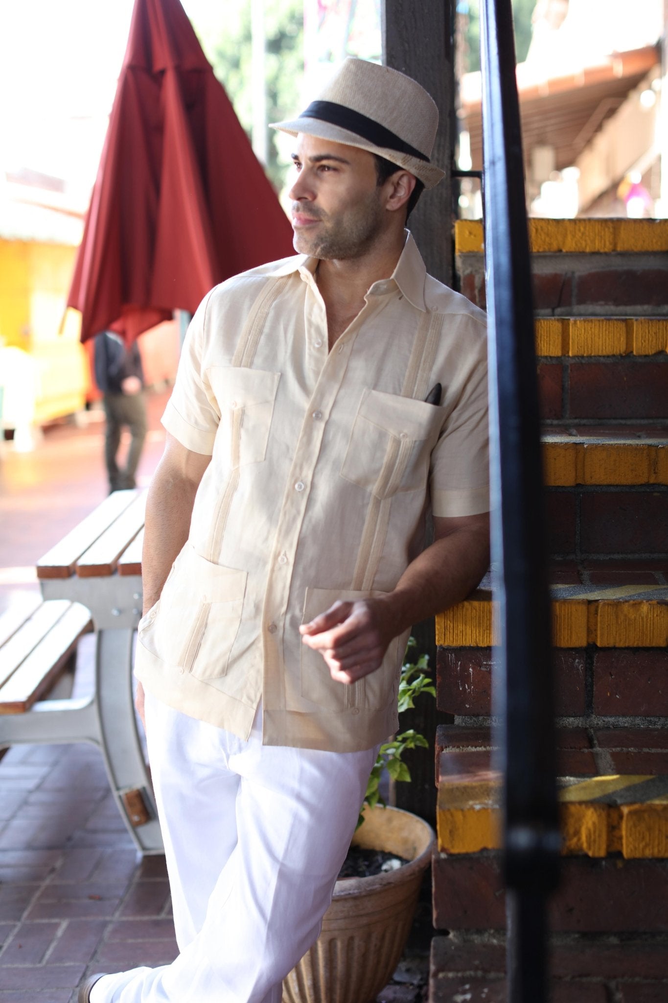 Man wearing the tan linen Guayabera Shirt and white pants, leaning against brick steps.
