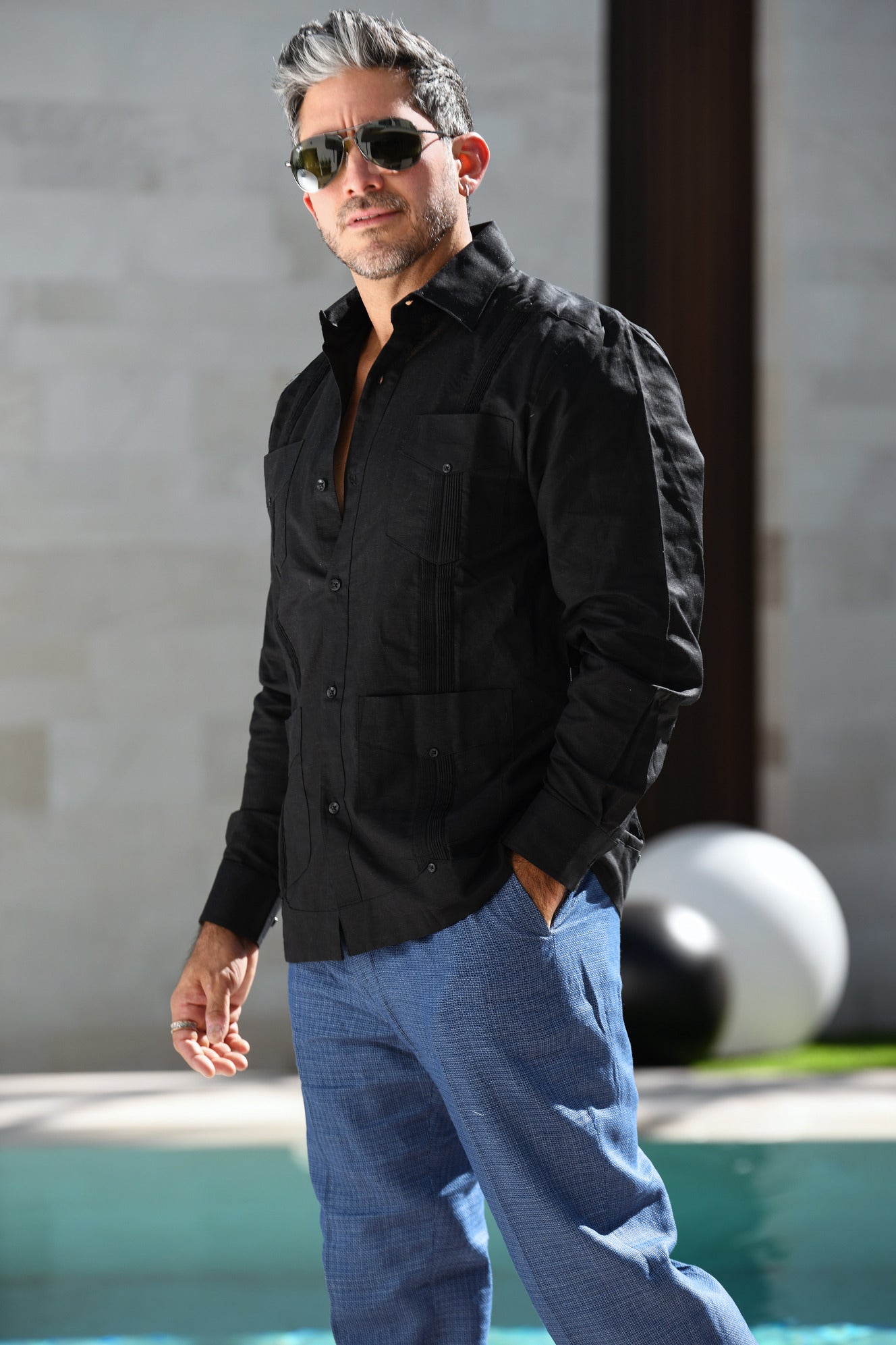 Man wearing a black guayabera and sunglasses standing by a poolside.