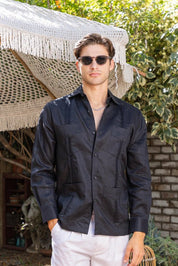 Man wearing sunglasses and a dark guayabera shirt outdoors with greenery and a woven chair in the background