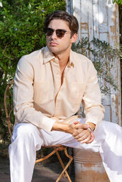 A man in a tan Men's Linen Classic Four-Pocket Long Sleeve Guayabera Shirt, seated outside.
