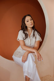 A woman sits wearing a beige Women's Short Sleeve Guayabera with 4 Pockets, with white pants.