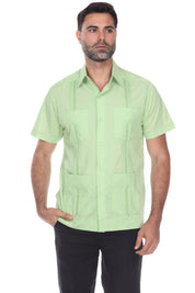 A man wearing a light green short-sleeve guayabera-style button-up shirt with four front pockets and black pants, standing against a plain white background.