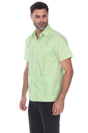 A man wearing a light green short-sleeve Guayabera-style shirt with front chest and waist pockets, paired with dark pants, photographed against a plain white studio background.
