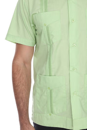 A close-up of a light green short-sleeve guayabera button-up shirt with multiple front pockets, worn by a model. The photo is cropped to show the torso and arm against a plain white background.