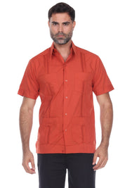 A man wearing a short-sleeve button-up shirt in a muted terracotta or burnt orange color, paired with black pants, photographed against a plain white background.