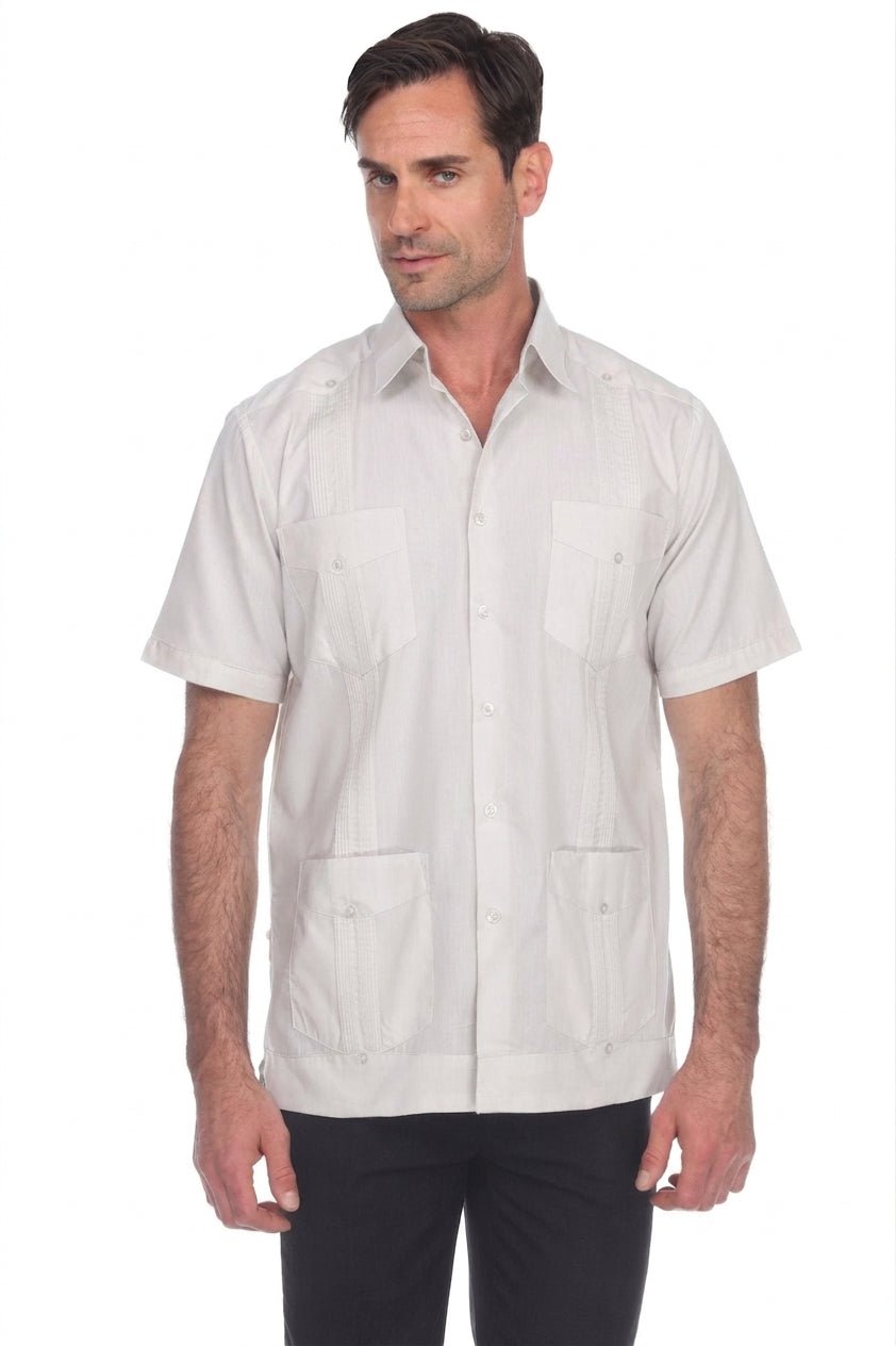A man wearing a light beige short-sleeve Guayabera-style button-up shirt with four front pockets, paired with black pants, photographed against a plain white background.
