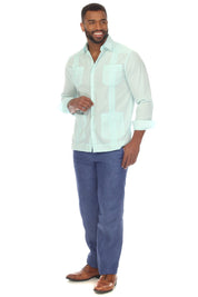 A man modeling a light mint green Guayabera-style button-down shirt with embroidered details, paired with blue pants and brown shoes, against a plain white background.