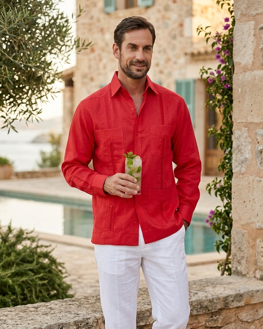 Man in a red guayabera shirt and white pants standing outdoors near a stone wall and building.